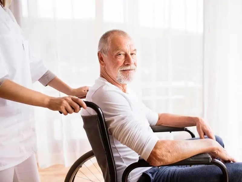 A carer in a white uniform pushes a smiling elderly man seated in a wheelchair by a bright window.