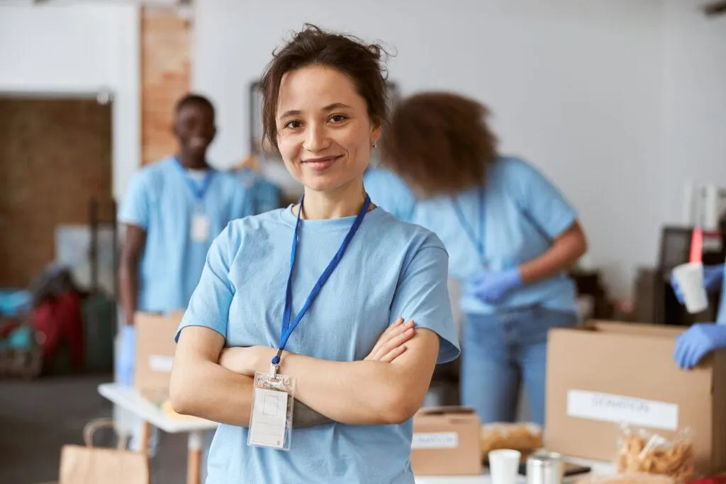 Smiling woman in a blue T-shirt with a lanyard stands with arms crossed as colleagues pack boxes behind her.