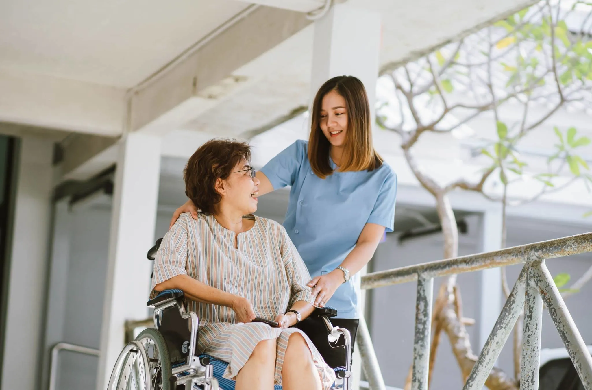 A young carer in a blue uniform smiles while assisting an older woman seated in a wheelchair on an outdoor ramp.